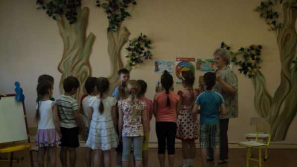 Воспитатель и дети стоят на фоне стены с изображением деревьев