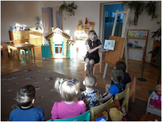 Воспитательница в тёмном коротком платье показывает детям картинки в книжке