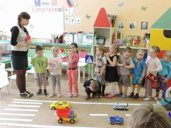 Воспитательница что-то говорит детям, стоящим перед макетом дороги в помещении группы