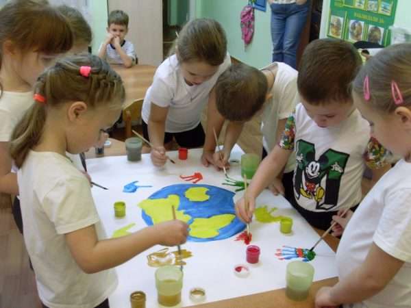 Дети рисуют красками коллективный плакат с изображением земного шара и цветными ладошками