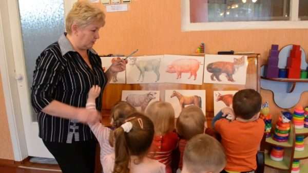 Воспитательница показывает детям картинки с изображениями животных на стенде