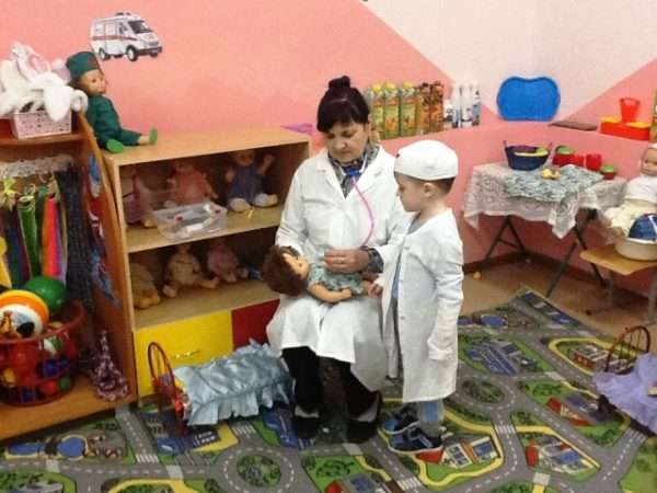 Воспитательница в белом халате объясняет мальчику, как пользоваться фонендоскопом