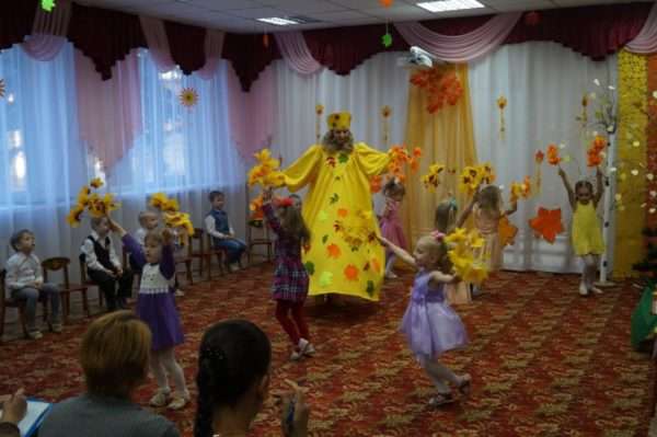 Дети и педагог в жёлтом костюме танцуют с осенними листьми