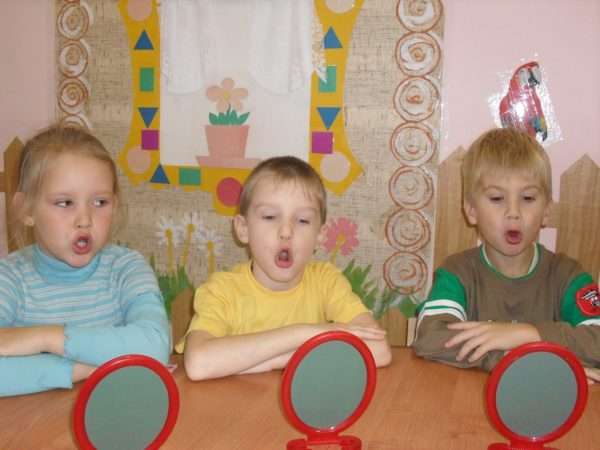 Трое детей выполняют артикуляционное упражнение, перед ними на столе стоят небольшие зеркала