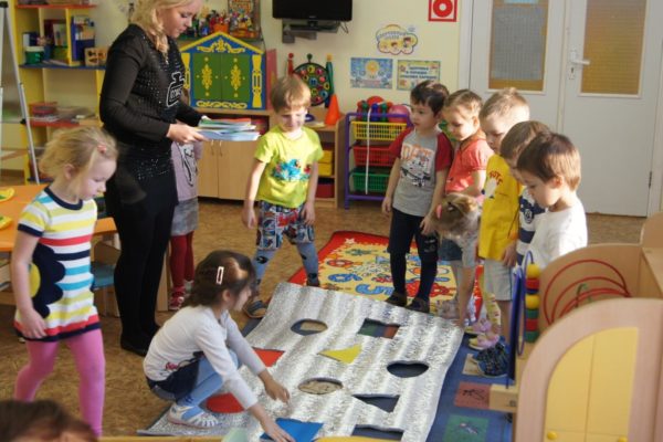 Дети и воспитатель стоят вокруг полотна с вырезанными геометрическими фигурами