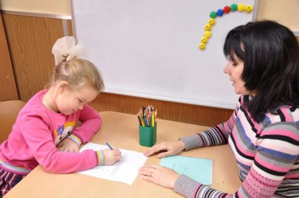 Девочка выполняет задание на листе бумаги, педагог что-то говорит