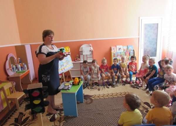 Воспитательница держит машинку и что-то рассказывает детям, сидящим полукругом
