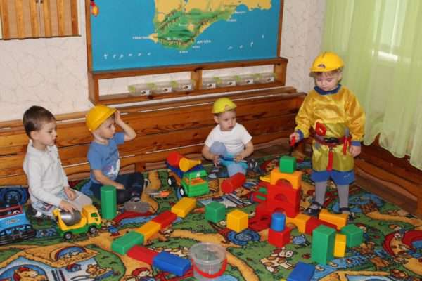 Четыре мальчика в жёлтых касках играют с машинками и строительным материалом