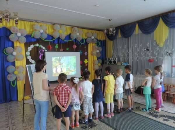 Воспитатель и дети стоят перед экраном, на котором демонстрируется мультфильм