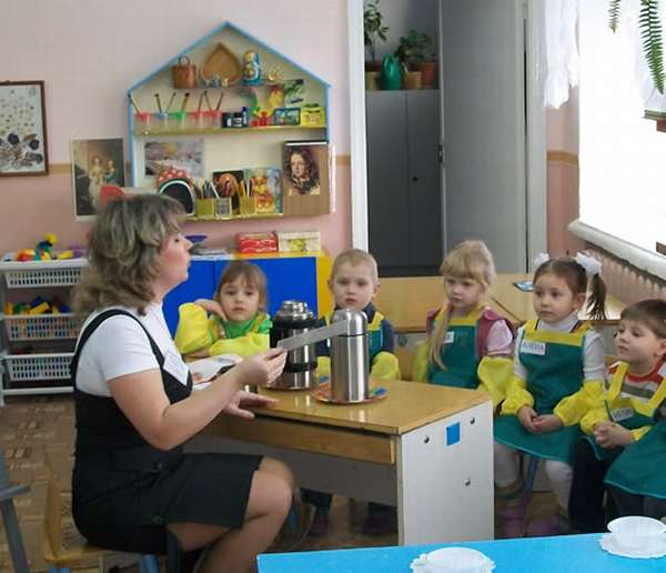 Воспитательница что-то рассказывает сидящим на стульях детям в зелёных фартуках